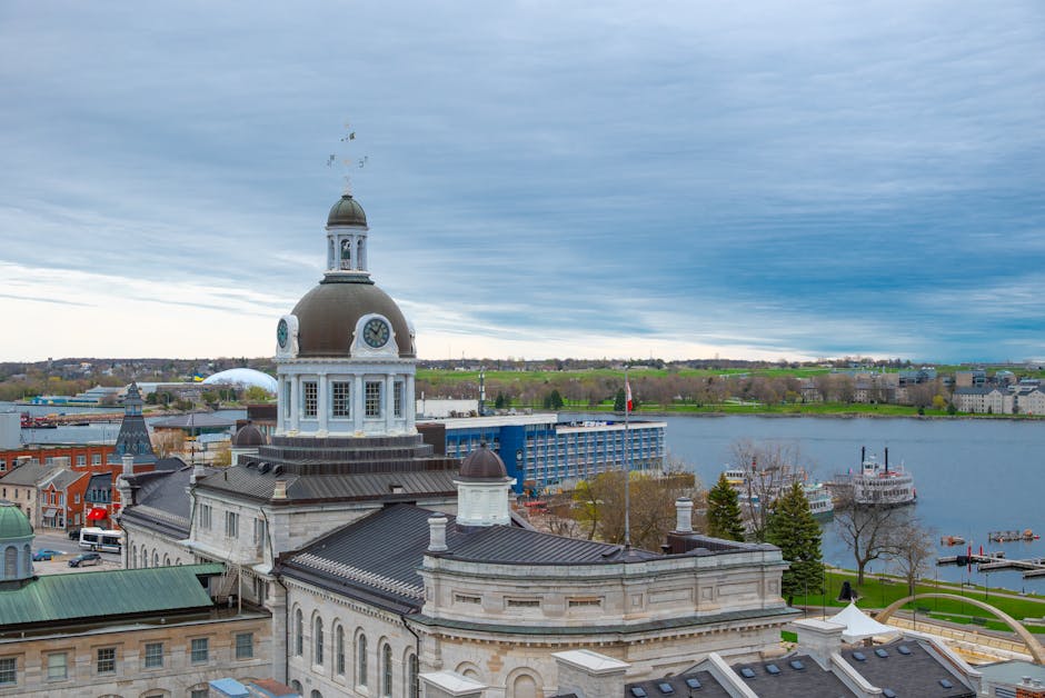 An aerial view of a historic building with a prominent clock tower featuring a domed roof, situated along the riverbank of Kingston. The scene includes a mix of architectural styles with classic stone structures and modern elements. The building's surface appears well-maintained and clean, with a smooth, dark roofing material. Surrounding areas include other residential and commercial buildings, some with green rooftops, as well as the river with boats docked along the waterfront. The sky is partly cloudy with diffused sunlight illuminating the scene, showcasing a serene urban landscape. This image exemplifies the importance of surface cleaning and maintenance within historic and modern architectural settings, as performed by companies like Deep Cleaning Kingston, who specialize in deep cleaning and sanitisation projects such as those on Clarence Street Kingston.