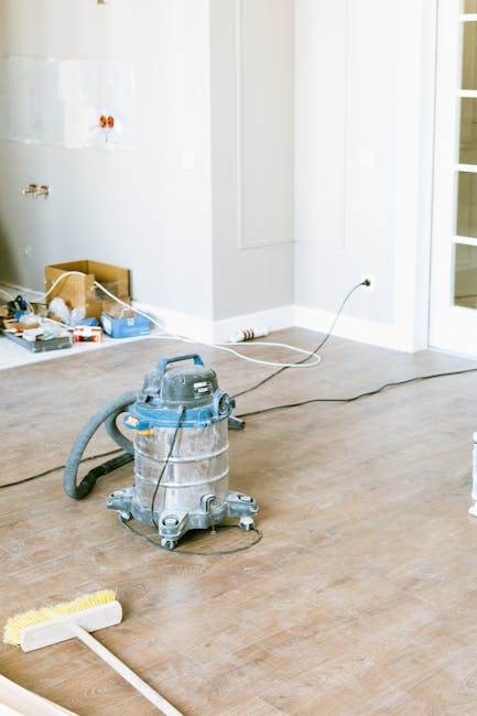 An unfinished room with light-colored wooden flooring undergoing surface cleaning as part of an after builders cleaning project. A commercial-grade vacuum cleaner is placed on the floor, with its hose extending toward the wall, which has visible plumbing fixtures and some construction tools and materials scattered in the corner. The room is well-lit by natural light coming through a glass door on the right. The walls are painted white, and the overall scene reflects a process of thorough cleaning and preparation, typical of services provided by Deep Cleaning Kingston for post-construction or renovation projects, ensuring a hygienic and polished finish.