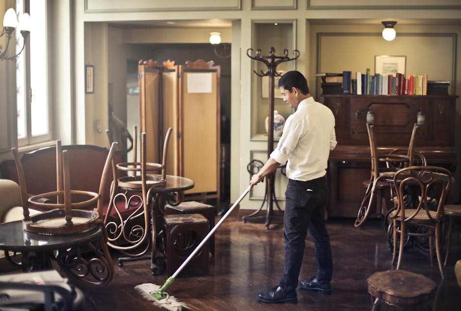 A professional cleaner from Deep Cleaning Kingston is using a mop to clean a dark wooden floor in a well-lit, traditional living room. The room features a collection of wooden chairs stacked on a roun