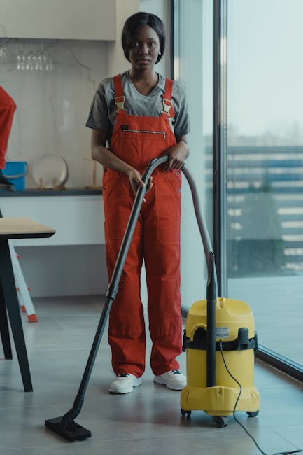 A professional cleaner from Deep Cleaning Kingston is using a mop to clean a dark wooden floor in a well-lit, traditional living room. The room features a collection of wooden chairs stacked on a roun