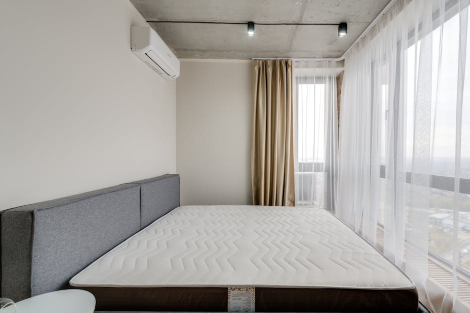 A modern bedroom featuring a neatly made bed with a dark gray upholstered headboard and a clean, white mattress. The room has large floor-to-ceiling windows covered with sheer white curtains and beige blackout drapes, allowing natural light to illuminate the space. The walls are painted in a neutral color and the ceiling is exposed concrete with recessed lighting. An air conditioning unit is mounted on the wall above the headboard, and the room appears tidy and well-maintained, reflecting professional deep cleaning standards by Deep Cleaning Kingston for surface cleaning, sanitisation, and domestic cleaning.