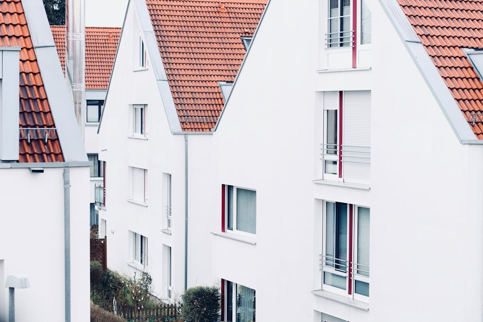 An exterior view of a row of modern residential buildings with white facades and red tiled pitched roofs. The buildings feature large windows, some with white shutters, and small balconies with metal railings. The scene is well-lit with natural daylight, highlighting the clean and freshly maintained appearance of the properties. This image exemplifies well-kept residential architecture, which may be part of a property estate serviced by Deep Cleaning Kingston for end of tenancy cleaning or general maintenance to uphold hygiene and appearance. The overall setting reflects a tidy, well-maintained housing area, suitable for domestic cleaning services. In the foreground, neatly trimmed shrubs and a garden fence are visible, contributing to the orderly aesthetic of the neighborhood.