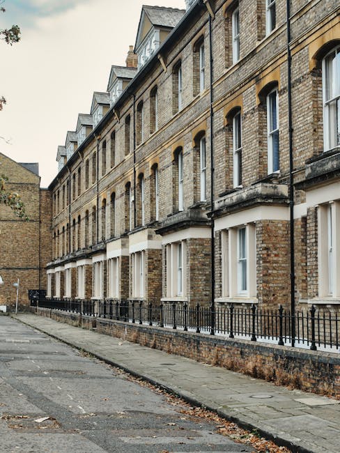 The image depicts a row of traditional brick residential buildings with large sash windows on each floor, set along a paved sidewalk. The buildings feature beige and brown bricks, with some architectural detailing around the windows and rooflines. Black iron railings run along the front of the buildings, separating the properties from the pedestrian walkway. The pavement appears clean, with no visible litter or debris, and the scene is illuminated by natural daylight, highlighting the texture of the brick surfaces. This setting exemplifies a typical urban residential area where surface cleaning and maintenance by professional services like Deep Cleaning Kingston are essential for preserving hygiene and visual appeal. The overall scene suggests a well-kept environment that benefits from regular deep cleaning, sanitisation, and domestic cleaning to maintain its appearance and hygiene standards, especially for tenant move-outs or end of tenancy cleaning for Norbiton estates and flats, Kingston.