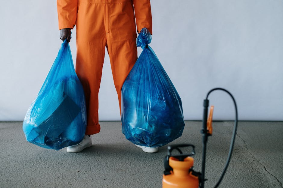 A person dressed in full personal protective equipment, including a white hazmat suit, safety goggles, and a respirator mask, standing amidst a large pile of mixed domestic and hazardous waste materia