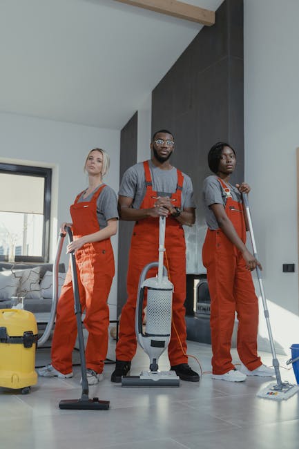 Three professional cleaners from Deep Cleaning Kingston, wearing grey shirts and bright orange overalls, are positioned in a modern living room with natural daylight coming through a window. They are each holding cleaning tools: one with a handheld vacuum, another with a spray bottle and cloth, and the third with a mop. The room features a polished wood floor, a glass coffee table, and a light-colored sofa in the background, all appearing well-maintained and clean. The bright, spacious room has a sleek, contemporary design with an emphasis on cleanliness and hygiene, reflecting deep cleaning and surface sanitisation processes typical of domestic cleaning services. Visible cleaning equipment includes a yellow vacuum cleaner, a spray bottle, and mop, indicating thorough surface cleaning and sanitisation. Deep Cleaning Kingston specializes in detailed, professional cleaning for residential spaces, ensuring surfaces such as floors, countertops, and glass are spotless and hygienic.