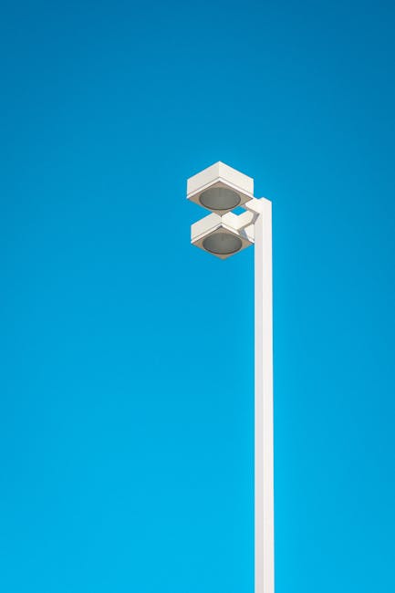 A tall, white street lamp with a modern, rectangular design stands against a clear blue sky. The lamp features two large, round lighting fixtures housed within a minimalistic enclosure at the top of the pole. The bright sunlight creates subtle shadows on the structure, emphasizing its clean, smooth surfaces. The scene appears to be outdoors in an urban or residential area, conveying a sense of cleanliness and maintenance associated with surface cleaning and illumination upkeep, as discussed in the context of hidden charges in Kingston cleaning quotes, with Deep Cleaning Kingston specializing in cleaning and sanitisation services.