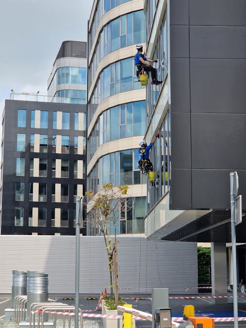 Two window cleaners operating high-rise scaffolding platforms on the exterior glass facade of a modern office building in Kingston. They are wearing safety helmets and harnesses, and are using cleaning tools and buckets to perform surface cleaning and window sanitisation. The building features large curved glass windows with reflective surfaces, set against a backdrop of other contemporary office structures with glass and steel elements. The street below includes a small tree, parking barriers, and safety tape, indicating active maintenance activity. This scene illustrates professional commercial cleaning services provided by Deep Cleaning Kingston, emphasizing high-level glass and surface cleaning for office environments.