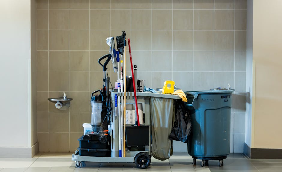 An organised cleaning trolley positioned against a beige tiled wall in an office cleaning setting, featuring various cleaning tools and supplies including a mop, broom, cloths, spray bottles, and a small vacuum cleaner. To the right, there is a large grey waste bin with a lid, and to the left, a small wall-mounted sink with a chrome faucet. The scene is well-lit, emphasizing the cleanliness and readiness for surface cleaning, disinfection, and deep cleaning processes. The image illustrates equipment used by Deep Cleaning Kingston for maintaining hygiene standards in office environments near Riverside Kingston and Canbury Gardens, Kingston.